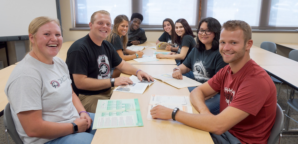 A group of students sitting all around a table smiling.